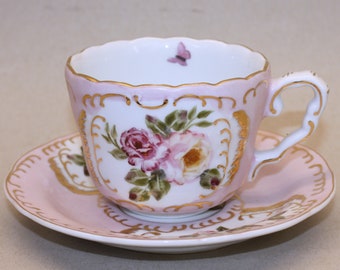 Könnte beinhalten: Ein rosa Teetassen- und Untertassen-Set mit Blumenmuster. Die Tasse hat einen goldenen Rand und ein zartes Schmetterlingsdesign. Die Untertasse hat ein passendes Blumenmuster und einen goldenen Rand.