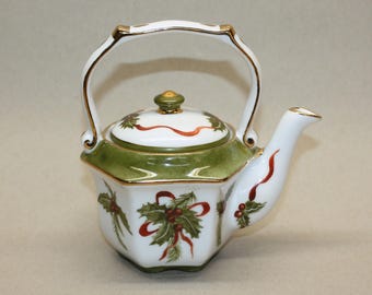 May include: White ceramic teapot with a green and gold trim, featuring holly and ribbon designs. The teapot has a curved handle and a lid with a gold knob. The teapot is a festive holiday item.