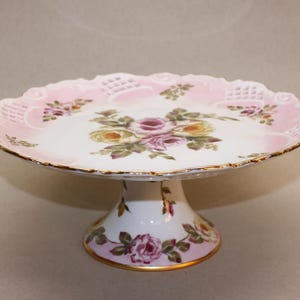 Puede incluir: Un soporte para tartas blanco con un diseño floral rosa y blanco. El soporte tiene un borde festoneado y un ribete dorado. La base del soporte también está decorada con flores.