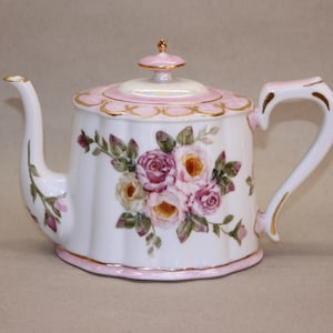 Puede incluir: Tetera de porcelana blanca con tapa y asa rosas, adornada con un diseño floral de rosas rosas y amarillas y hojas verdes. La tetera tiene ribete dorado y un pico curvo, ideal para servir té.