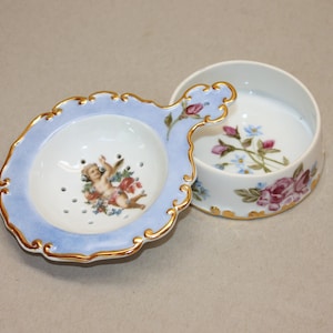 May include: A porcelain tea strainer and matching bowl set featuring a light blue border with gold trim. The strainer depicts a cherub, while the bowl features a floral motif with pink roses and blue flowers painted on the white ceramic surface.