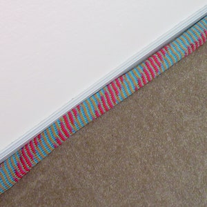 May include: A colorful door draft stopper with a knitted design. The stopper features a pattern of pink, blue, and gold stripes. A wooden button is attached to one end. The item is placed against a white wall and a carpeted floor.