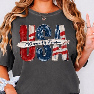 Peut inclure: T-shirt gris fonc&eacute; avec un motif patriotique "USA". Les lettres "USA" sont rouges, blanches et bleues, avec le texte "250 years Of Freedom" au centre. Le t-shirt a des manches courtes et un col rond.