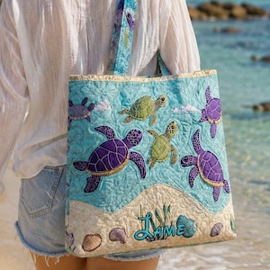 May include: A quilted tote bag with a light blue background featuring embroidered sea turtles in shades of green and purple. The bag has a sandy beach design at the bottom with the word "Lame" embroidered in a script font. The bag is carried by a person on a beach.