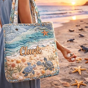 May include: A quilted tote bag with a beach scene design. The bag features a blue ocean, sand, seashells, starfish, and two sea turtles. The name "Clark" is embroidered on the front. The bag has a patterned strap and is carried on a beach.