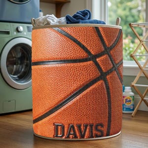 May include: A cylindrical laundry hamper with a basketball design. The hamper is orange with black lines and the name "DAVIS" in black lettering. The hamper is filled with folded clothes and has beige handles.
