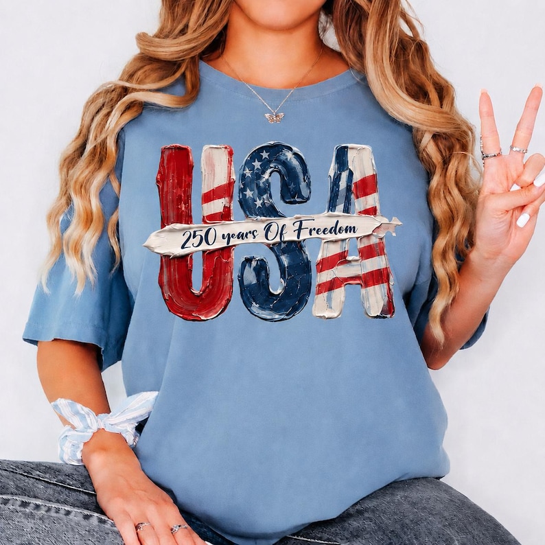 Peut inclure: T-shirt bleu clair avec le mot "USA" en rouge, blanc et bleu, rappelant le drapeau am&eacute;ricain. Le texte "250 years Of Freedom" est imprim&eacute; au centre. Le t-shirt a une coupe d&eacute;contract&eacute;e.