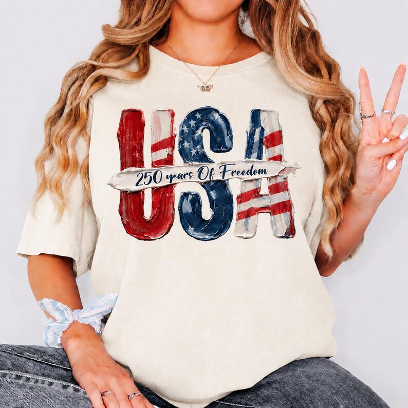 Peut inclure: T-shirt cr&egrave;me avec le mot "USA" en rouge, blanc et bleu, avec un motif de drapeau vieilli. Le texte "250 years Of Freedom" est imprim&eacute; en dessous du mot "USA".