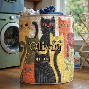 May include: A round laundry basket with a quilted fabric exterior. The fabric features a repeating pattern of colourful cats. The name "Olivia" is embroidered on the front. The basket has beige handles and is filled with laundry.