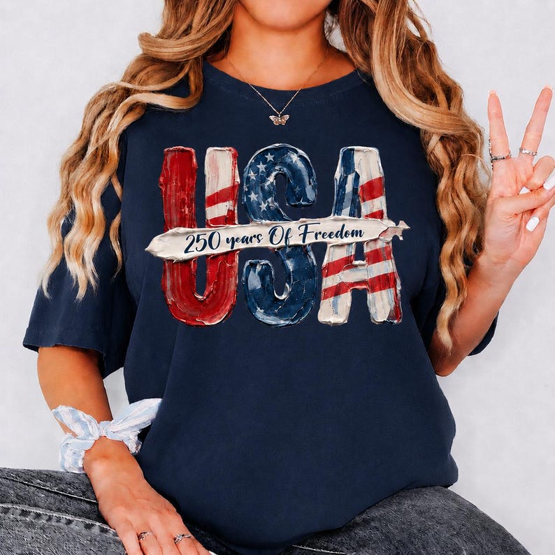 Peut inclure: T-shirt bleu marine avec un motif patriotique. Le mot "USA" est affich&eacute; en rouge, blanc et bleu, avec un effet drapeau vieilli. Le texte "250 years Of Freedom" est centr&eacute; en dessous. Un t-shirt d&eacute;contract&eacute; &agrave; col rond.