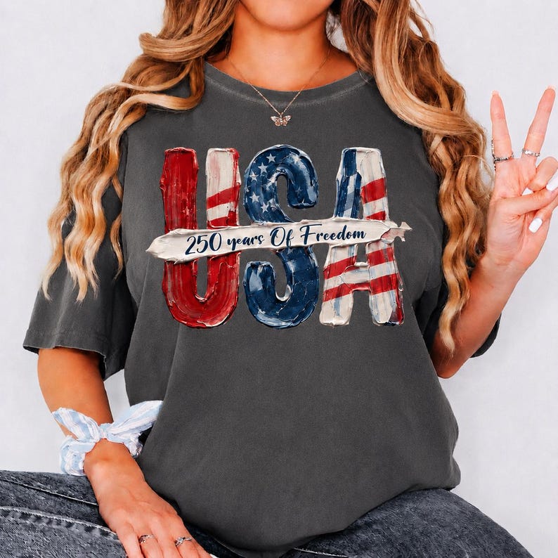 Peut inclure: T-shirt gris fonc&eacute; avec un motif patriotique "USA". Les lettres "USA" sont rouges, blanches et bleues, avec le texte "250 years Of Freedom" au centre. Le t-shirt a des manches courtes et un col rond.
