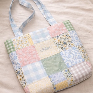 May include: A patchwork tote bag in pastel shades. The bag has a light blue gingham pattern on the handles and a floral pattern. The name "chloe" is embroidered on the front. The bag is made of various fabric squares.