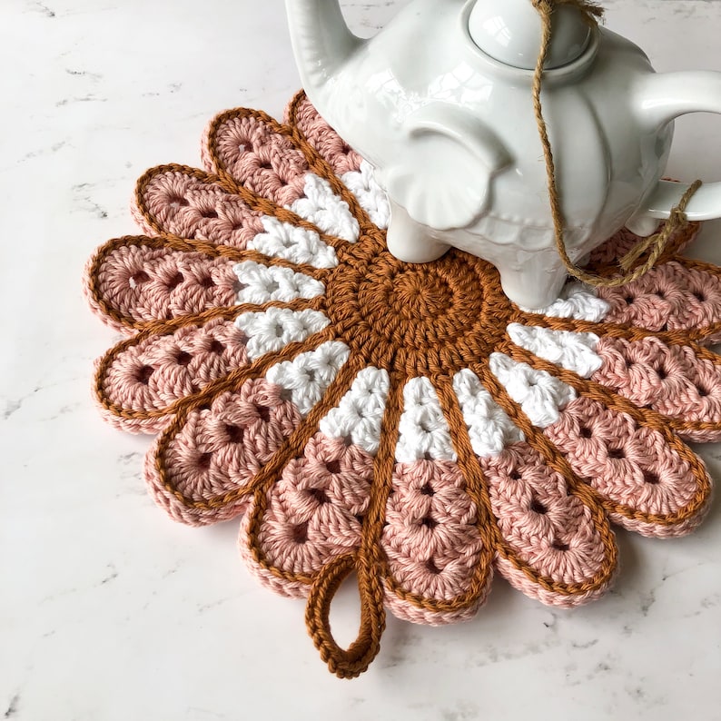 May include: A crocheted pot holder in the shape of a flower with pink, white, and brown yarn. The pot holder has a loop handle.