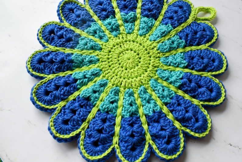 May include: A crocheted flower-shaped pot holder in shades of blue and green. The center of the pot holder is a green circle with a blue border. The outer edges of the pot holder are blue with a green border.