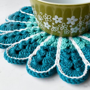 May include: A teal blue crocheted coaster with a white border, featuring a flower-like design. A green bowl with white floral pattern sits on top of the coaster.
