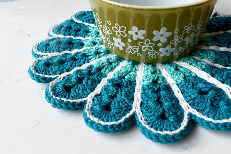 May include: A teal blue crocheted coaster with a white border, featuring a flower-like design. A green bowl with white floral pattern sits on top of the coaster.