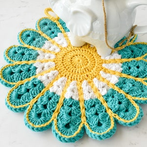 May include: A crocheted flower-shaped trivet in teal, white, and yellow. The trivet is a circular design with a center of yellow, then white, and then teal petals.