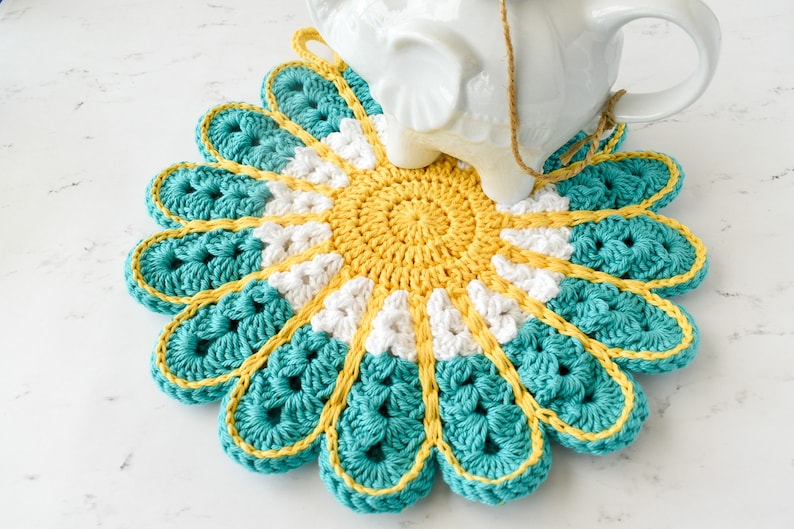 May include: A crocheted flower-shaped trivet in teal, white, and yellow. The trivet is a circular design with a center of yellow, then white, and then teal petals.