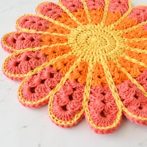 May include: A crocheted flower-shaped coaster in shades of pink, orange, and yellow. The coaster is made with a variety of stitches and has a textured surface.