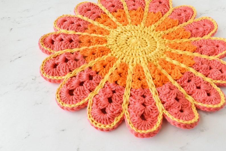 May include: A crocheted flower-shaped coaster in shades of pink, orange, and yellow. The coaster is made with a variety of stitches and has a textured surface.