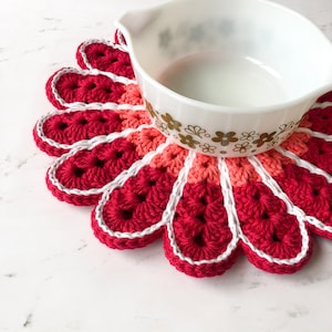 May include: A red, pink, and white crocheted flower-shaped hot pad with a white rim. A white ceramic bowl with a gold floral pattern sits on top of the hot pad.