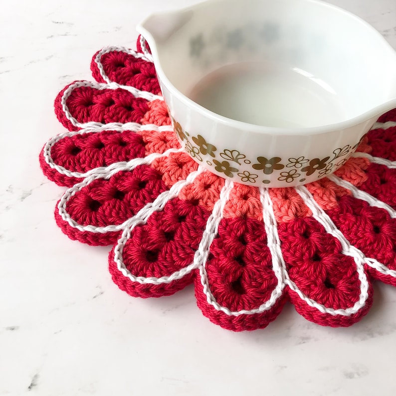 May include: A red, pink, and white crocheted flower-shaped hot pad with a white rim. A white ceramic bowl with a gold floral pattern sits on top of the hot pad.