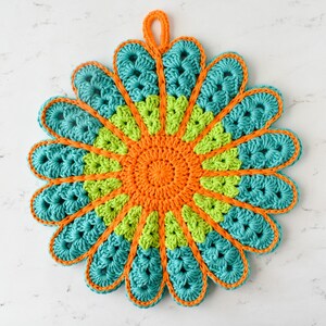May include: A crocheted trivet in the shape of a flower with teal, lime green, and orange yarn. The trivet has a loop for hanging.