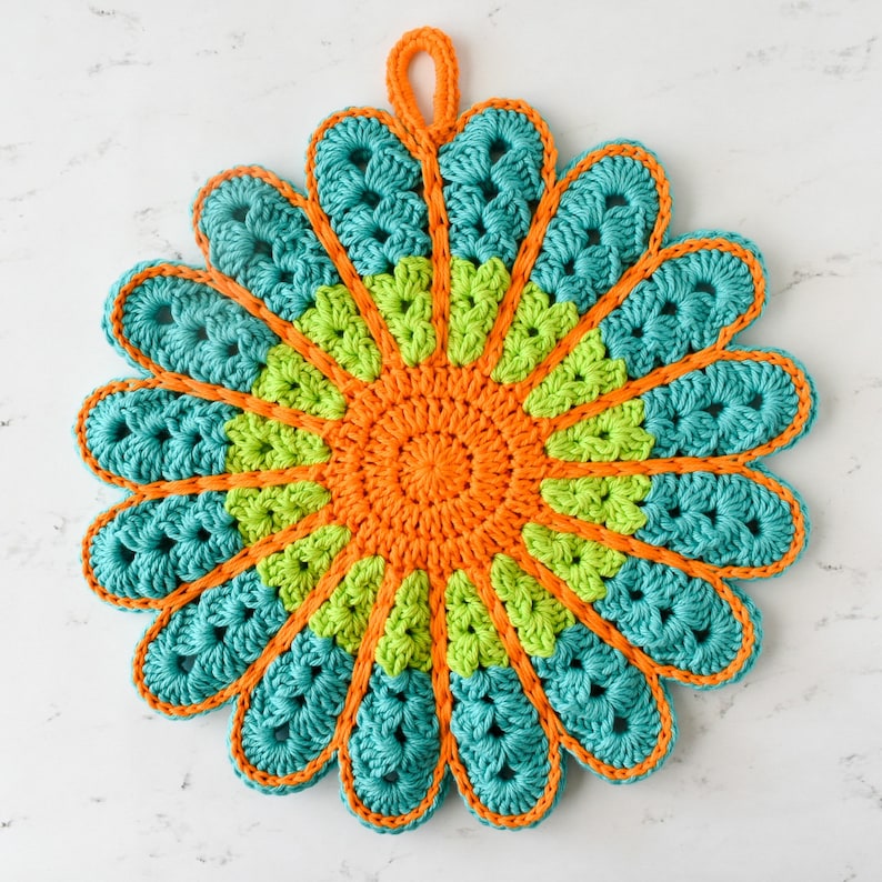 May include: A crocheted trivet in the shape of a flower with teal, lime green, and orange yarn. The trivet has a loop for hanging.