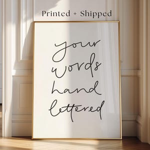 Può includere: Una stampa artistica rettangolare verticale in una sottile cornice dorata. Lo sfondo bianco presenta una scritta nera in corsivo che recita your words hand lettered. Sopra la cornice, il testo Printed + Shipped è chiaramente visibile.