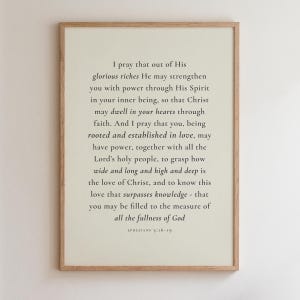 May include: A vertical rectangular wall art print in a light wood frame featuring black serif text on a cream background. The text displays the Bible passage Ephesians 3:16-19, beginning with 'I pray that out of His glorious riches' and ending with 'all the fullness of God'.