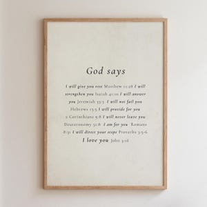 May include: A vertical rectangular wall art print in a light wood frame featuring black serif text on a cream background. The text reads: God says, followed by various biblical verses including Matthew 11:28, Isaiah 41:10, and John 3:16.