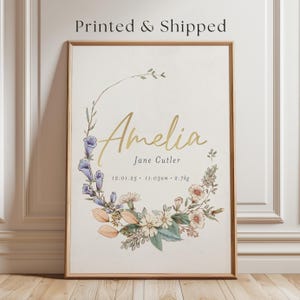 May include: A vertical rectangular wall art print in a wooden frame featuring the name Amelia and Jane Cutler in gold and black text. Below the name, the text reads 12.01.25, 11.03am, and 2.7kg. A floral wreath with blue and peach flowers surrounds the text.