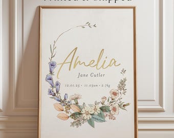Könnte beinhalten: Ein vertikales, rechteckiges Wandbild in einem Holzrahmen mit dem Namen Amelia und Jane Cutler in goldener und schwarzer Schrift. Unter dem Namen steht der Text 12.01.25, 11.03am und 2.7kg. Ein Blumenkranz mit blauen und pfirsichfarbenen Blüten umrahmt den Text.