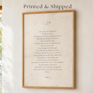 May include: A framed wall art print featuring the text of Psalm 23 in a dark serif font on a light beige, textured background. Above the text is a small, simple line drawing of a sprig. The frame is made of light-coloured wood.