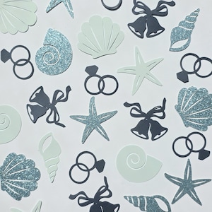 May include: Wedding-themed confetti featuring seashells, starfish, wedding bells, and engagement rings in light blue, glittery blue, and grey. The confetti is scattered on a white background, suitable for wedding decorations.