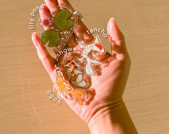 Könnte beinhalten: Transparente Koi-Teich-Schlüsselanhänger aus Acryl mit verschiedenen Koi-Fisch-Designs, darunter Showa, Tancho und Ogon, sowie einem Seerosen- und Blumendesign. Die Schlüsselanhänger werden in einer Hand gehalten, mit dem Text "koi pond keychains" unten.
