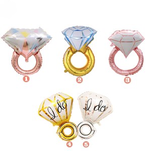May include: A collection of diamond ring-shaped balloons in various colors. The balloons include a rose gold ring with a white and pink diamond, a gold ring with a blue diamond, and a rose gold ring with a white and pink diamond. Two balloons say "I do".