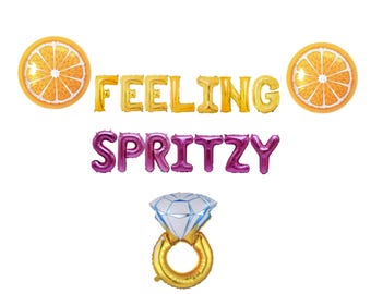 May include: Orange slice foil balloons spelling "Feeling Spritzy" with a gold ring and diamond balloon.