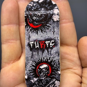May include: A fingerboard deck featuring a black, white, and red graphic illustration. The design includes a stylised skull with a red halo and the text "TURPS" in a bold, jagged font centred between a dark cloud and the skull figure.