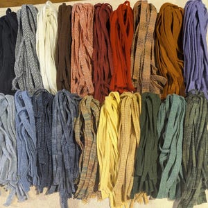 May include: A collection of fabric strips arranged in two rows, featuring a variety of solid colors and woven patterns including black, gray, white, brown, orange, red, tan, mustard, purple, light blue, navy, yellow, green, and teal.