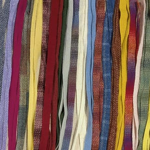 May include: A collection of long, narrow fabric strips arranged vertically in a side-by-side pattern. The strips feature a diverse array of colors including yellow, red, blue, green, purple, brown, and beige, with various textures and woven patterns.