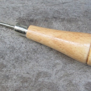 Puede incluir: Una herramienta de madera con una punta de metal, utilizada para perforar agujeros en cuero. La herramienta tiene un mango de madera y una punta de metal.