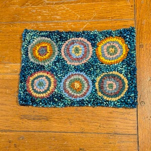 May include: Rectangular rug with six circular patterns in shades of blue, orange, yellow, and pink. The rug is handmade, with each circle featuring a different colour combination. The background is a speckled blue.