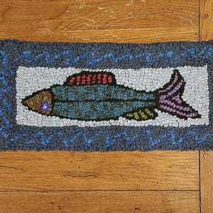 Puede incluir: Una alfombra de pescado azul, verde, rojo y morado con fondo blanco y borde azul. La alfombra está hecha de hilo y tiene una superficie texturizada.