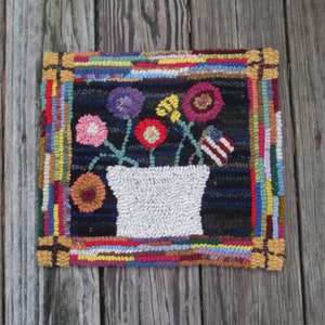 May include: A colourful rug with a white pot and flowers in the centre. The rug has a black background with a multi-coloured border.