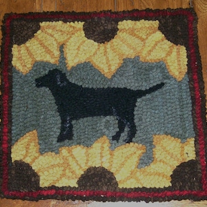 Op de afbeelding: Een zwart honden silhouet op een tapijt met een geel, bruin en grijs patroon. Het tapijt heeft een rode en bruine rand.