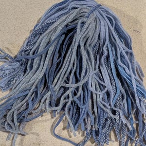 May include: A bundle of blue and gray dreadlocks, with varying shades and textures. Some strands are solid, while others have patterned details. The dreadlocks are arranged on a neutral-toned surface, suggesting a craft or hair accessory.
