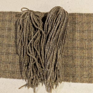 Peut inclure: Gros plan sur un tissu tweed marron et beige avec une bordure effilochée. Deux paquets de longs matériaux effilochés et fins sont posés sur le tissu. Le tissu présente un motif à carreaux.