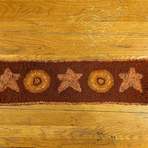 May include: A rectangular, brown rug with a repeating pattern of stars and circles in shades of orange and tan. The rug is placed on a wooden surface.