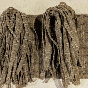 Peut inclure: Deux paquets de bandes de tissu marron avec une bordure effilochée, reposant sur un tissu à carreaux assorti. Le tissu a un aspect texturé avec un mélange de fils marron, beige et blancs, créant un motif à carreaux subtil. Les bandes sont nouées en haut.